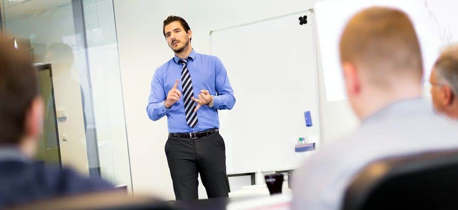 A young male project manager stands in front of a whiteboard, presenting at a team meeting.