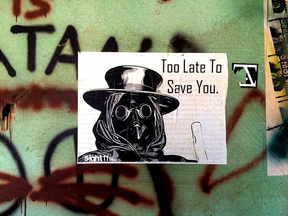 A black and white piece of art featuring a bird with goggles, a top hat and a hood, with the caption “Too Late to Save You,” stuck to a wall with spraypainted graffiti.