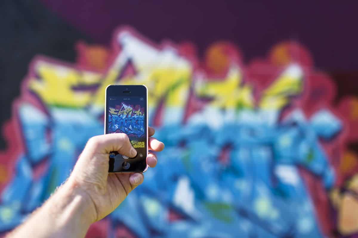 A close up of the hand of a young man holding a mobile phone and taking a picture of a blurred graffiti backdrop which is captured clearly on the screen.