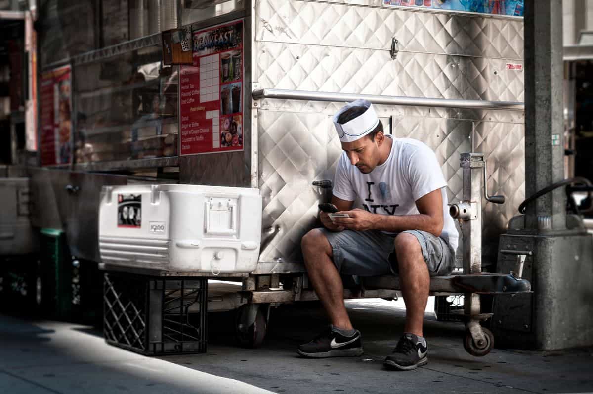 A young man wearing a cook's hat sitting next to a food truck in New York, comfortably busy on his mobile phone.