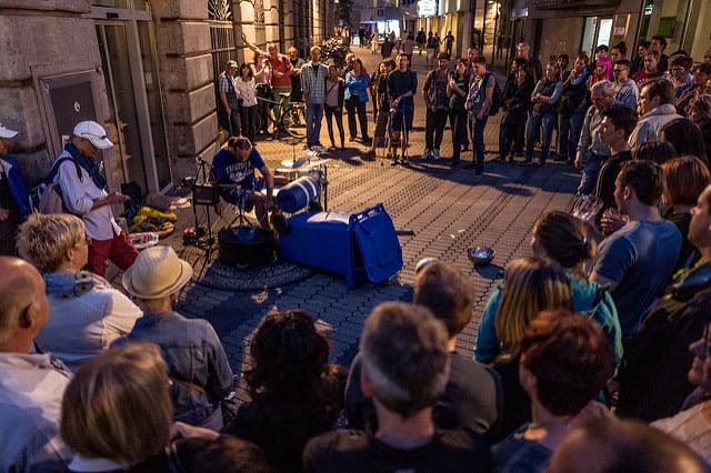 A photo of a crowd gathering to watch a street performer use unique pieces as a drum set.