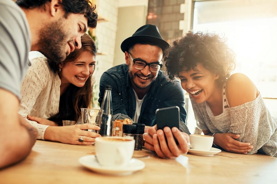 A photo of four people smiling and laughing gleefully at something on a smartphone.