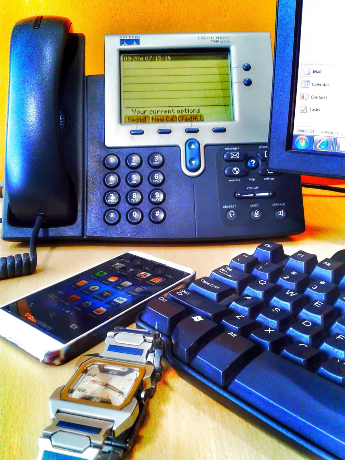 A smartphone, watch, computer and office phone on a desk.