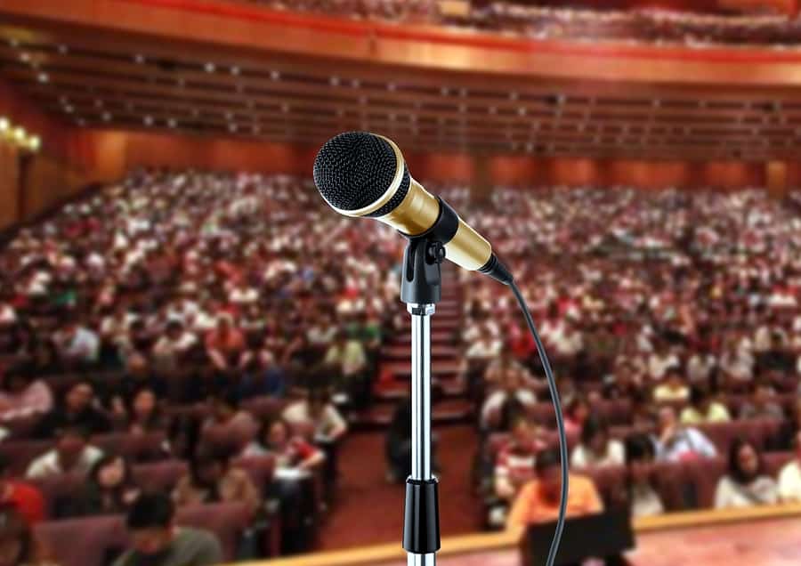 A close-up photo of a microphone on a mic stand with a large audience in the background.