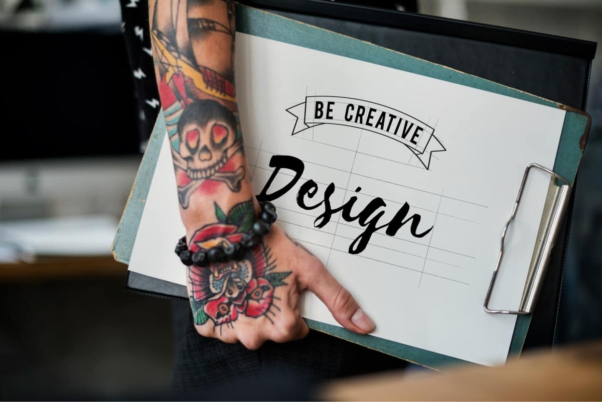 A person with tattoos on their arm holding a clipboard that says “be creative design.”