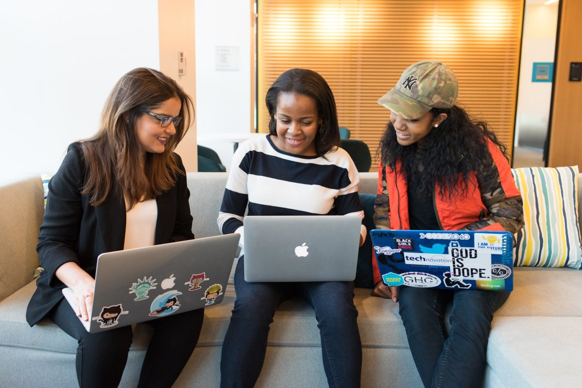 A photo of three women sitting next to each other on a couch conferring over a project, each with a laptop in their laps.
